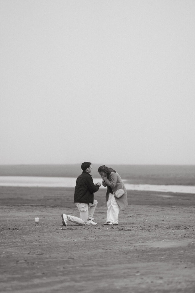 Man proposing on one knee on a quiet, misty beach as his partner reacts with emotion—perfect for ideas for engagement photos that feel deeply personal.