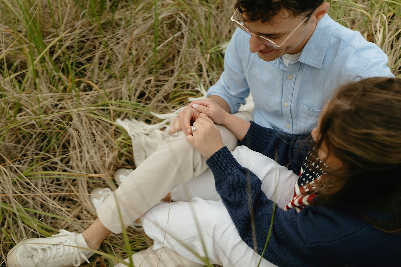 Couple sitting together in tall grass, admiring the engagement ring—an organic and heartfelt idea for engagement photos outdoors.