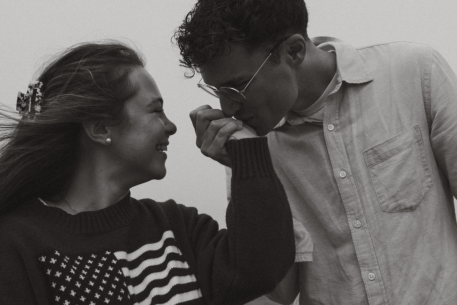 Black and white image of a man kissing his partner’s hand, both smiling softly, evoking timeless romance.