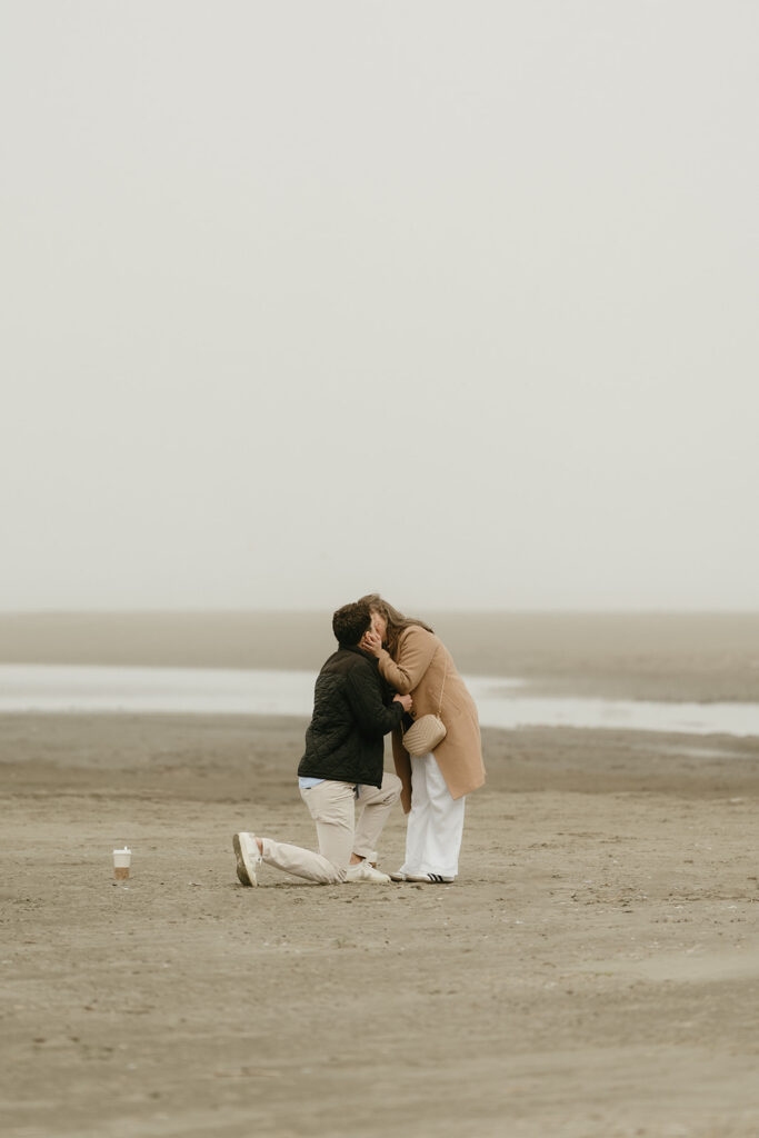 Couple kissing just after a beach proposal, surrounded by fog and shoreline.