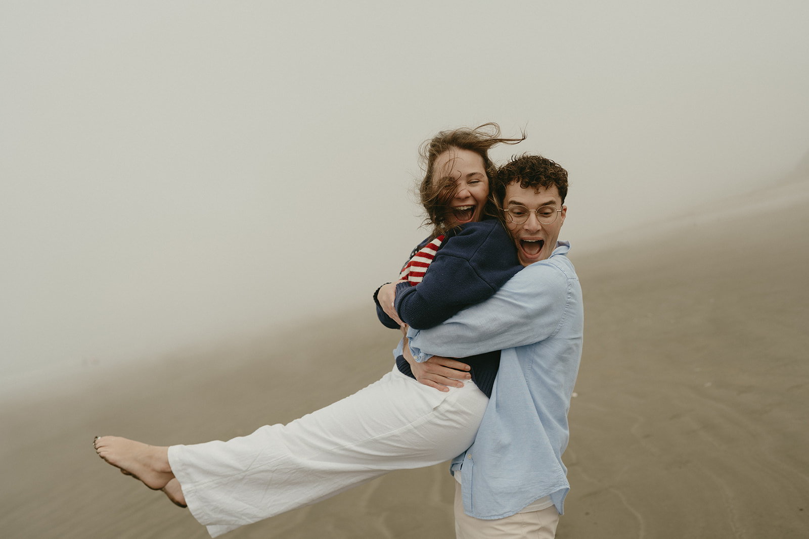 Playful moment on the foggy beach as the man lifts his fiancée, both laughing—ideal for couples wanting fun and lighthearted ideas for engagement photos.