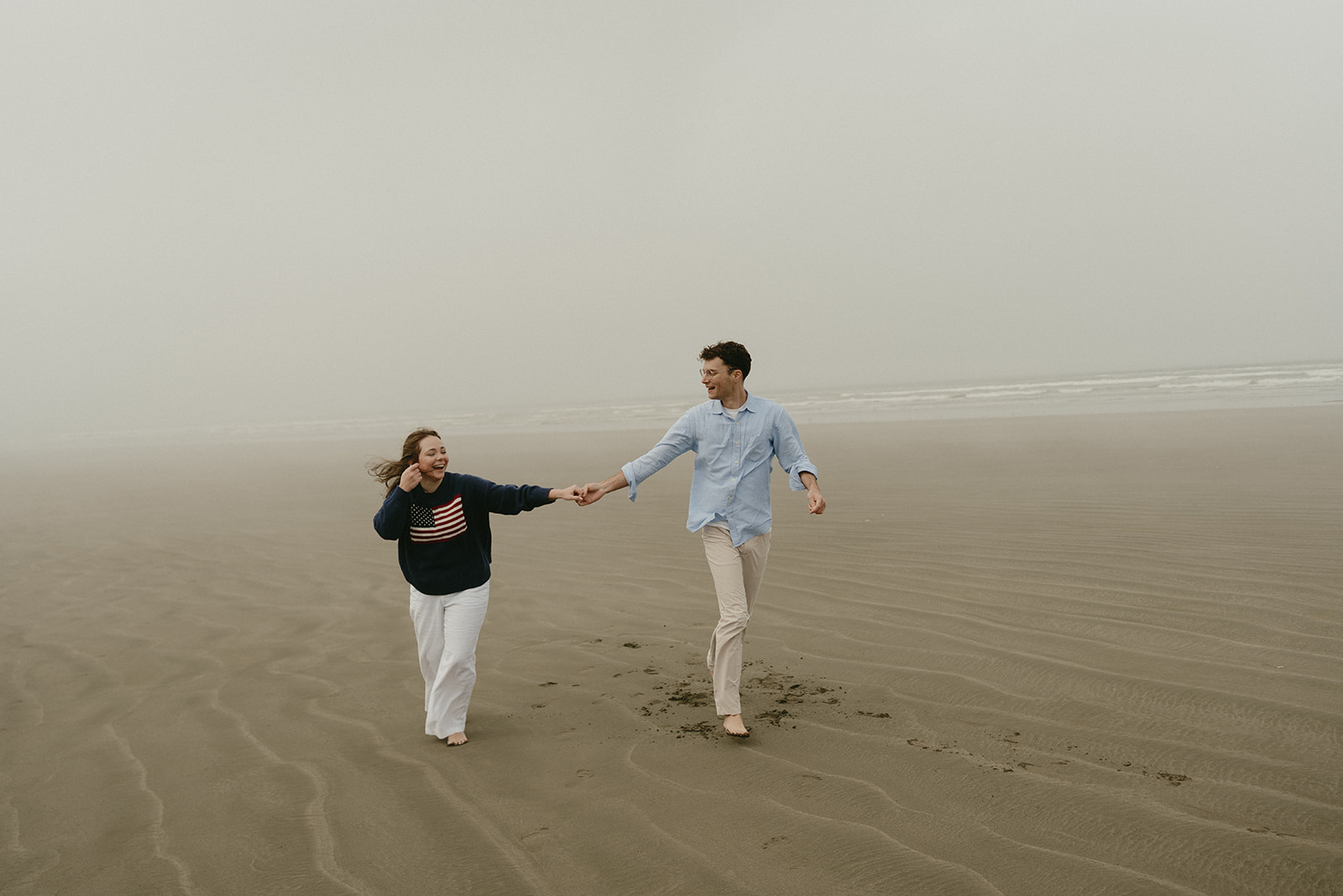 Couple running barefoot on the beach, holding hands and laughing, footprints trailing behind them.