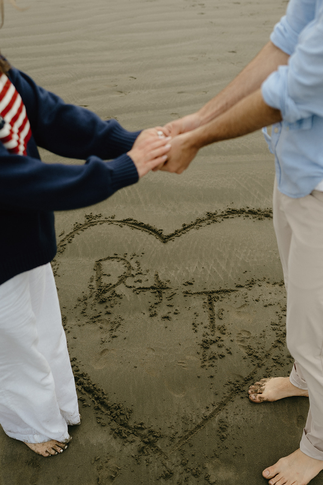 Couple holding hands above a heart drawn in the sand with their initials inside, a sweet and simple engagement detail.