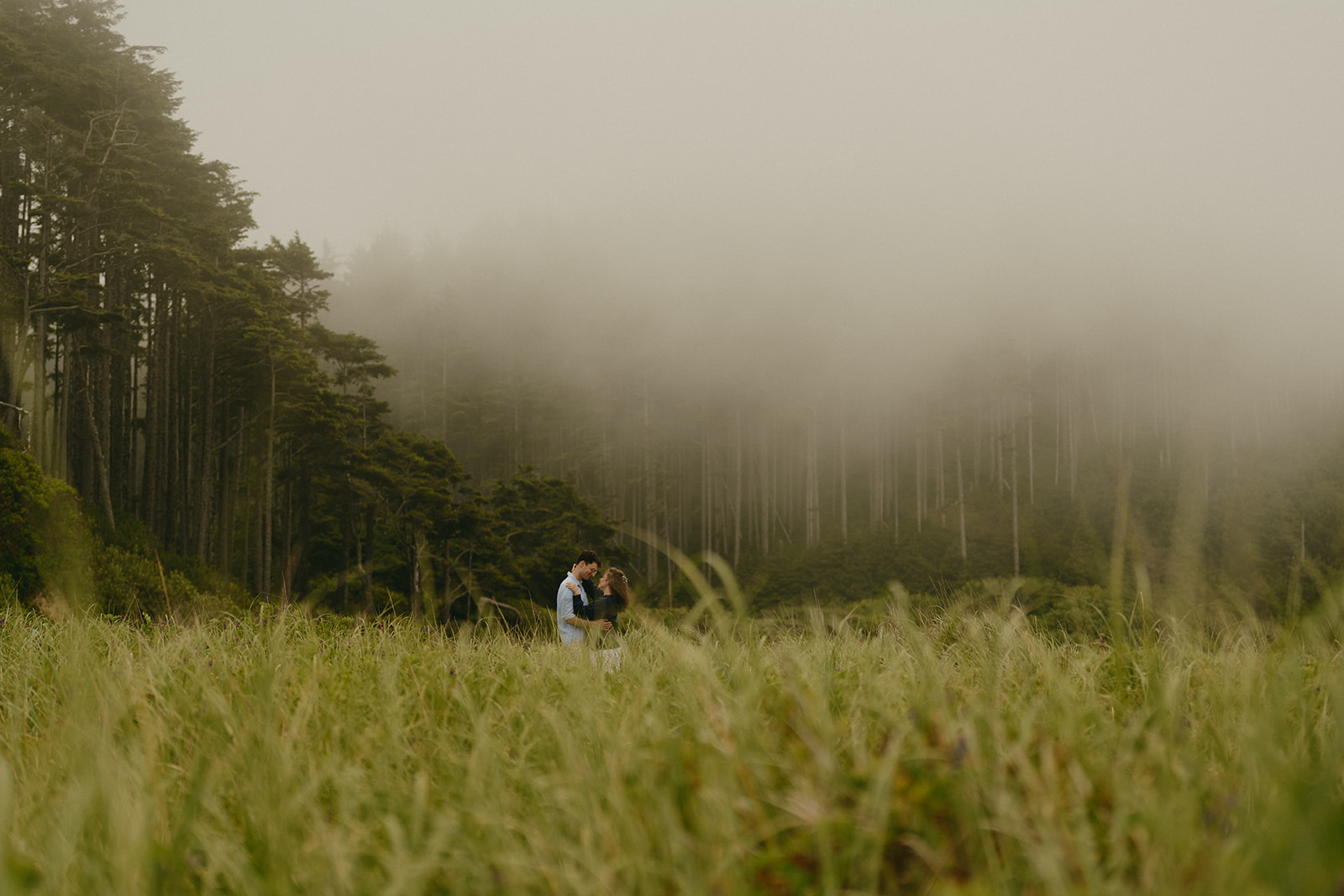 Couple embracing in a wide meadow beneath a mist-covered forest, a dreamy and cinematic location that offers inspiring ideas for engagement photos in the Pacific Northwest.