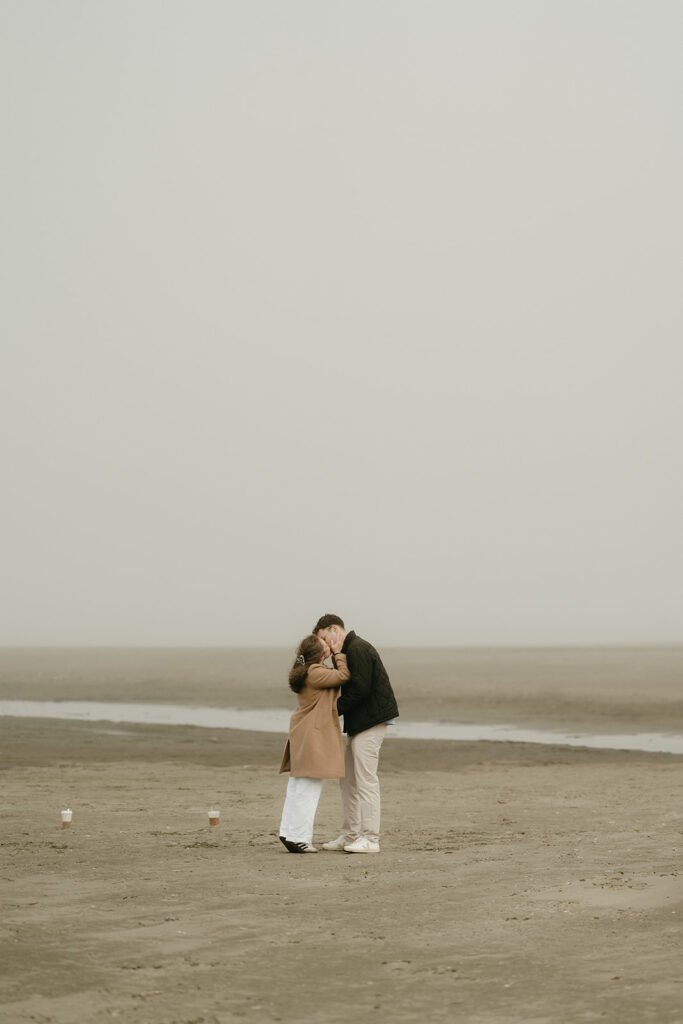 Couple sharing a soft kiss on a wide, empty beach with fog in the background.