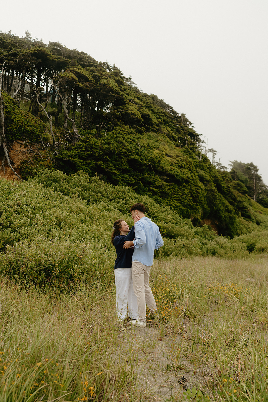 Couple sharing a lighthearted moment in a field of tall grass, surrounded by forested cliffs.