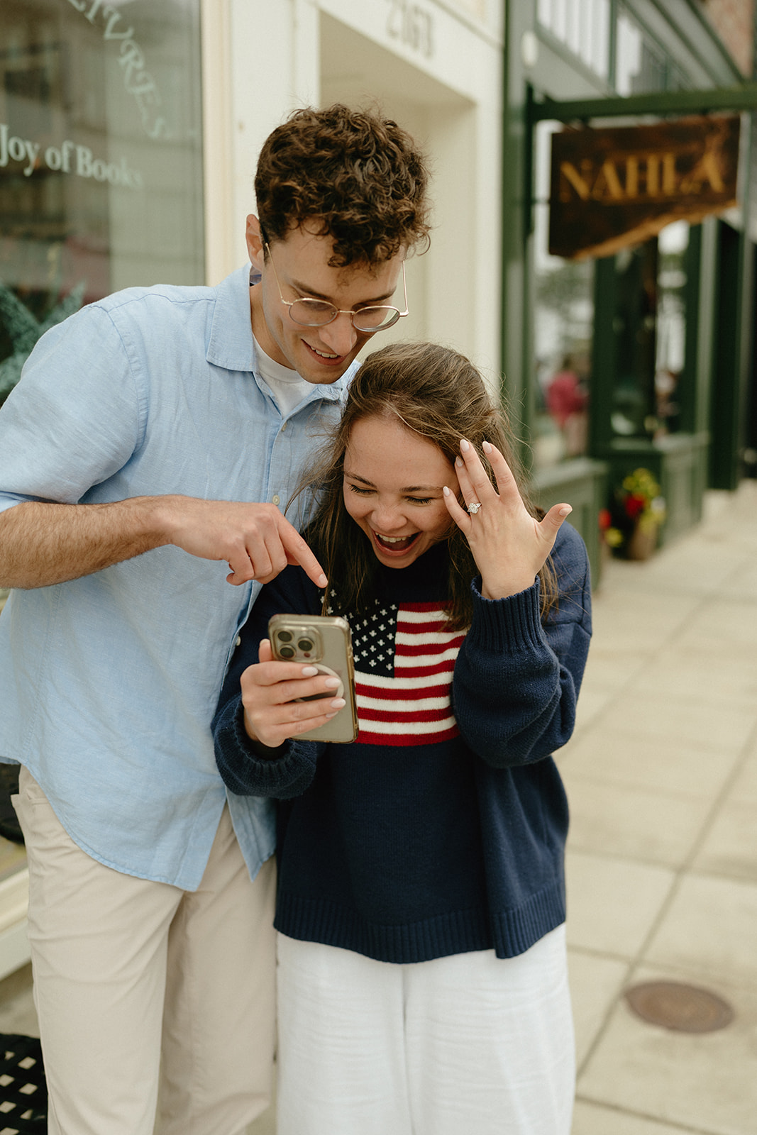 Couple laughing and pointing at a phone screen while celebrating their engagement in a small town setting.