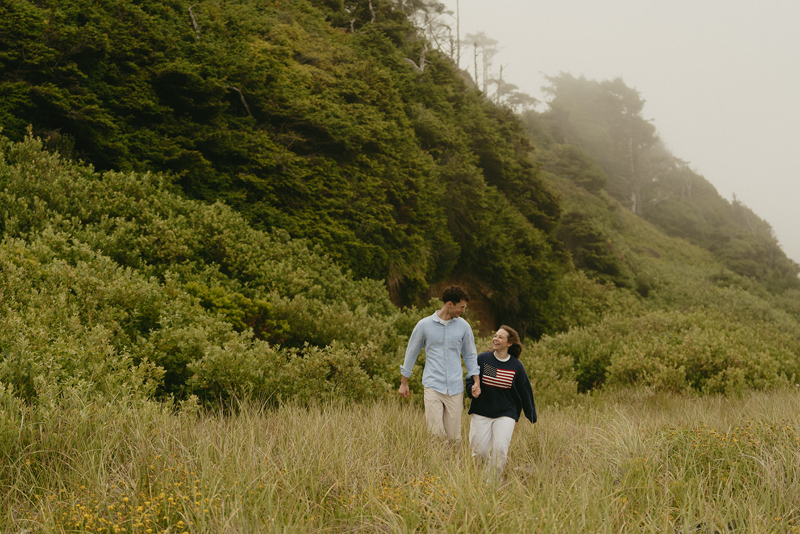 Couple walking hand in hand through a coastal meadow, sharing a quiet moment beneath misty skies.