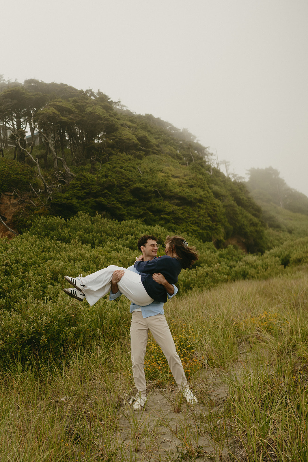 Man lifting his partner playfully in a misty coastal meadow, surrounded by lush greenery.