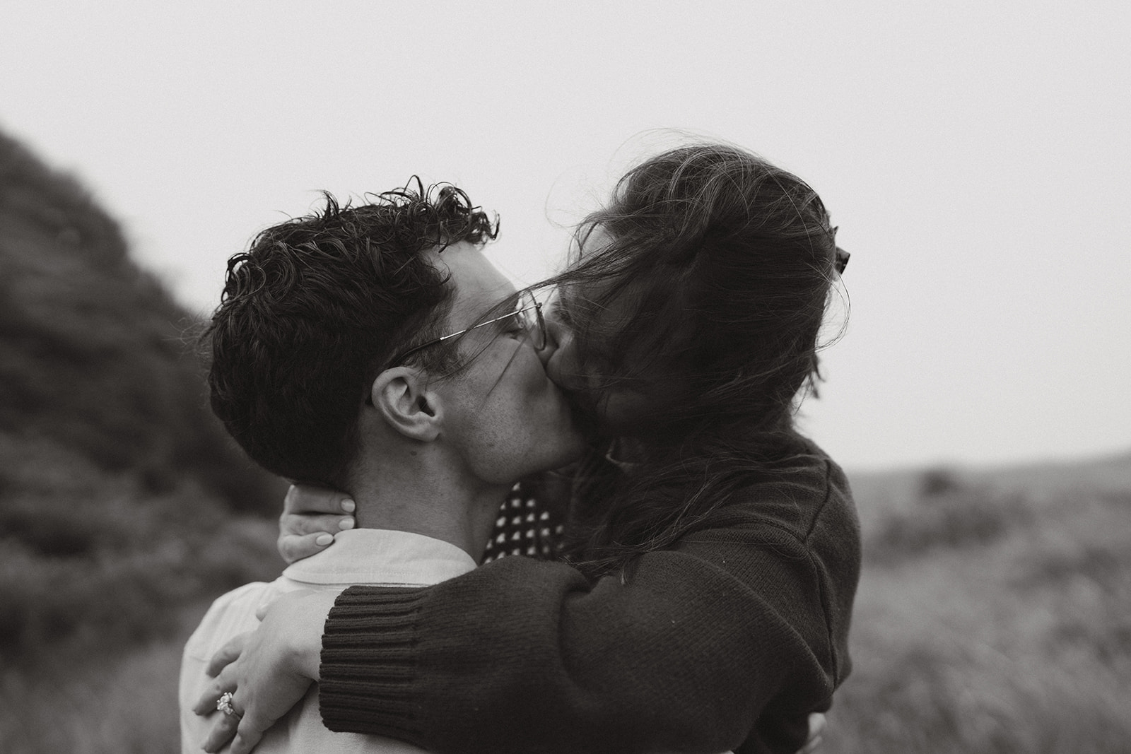 Black and white photo of a couple kissing, wind gently lifting her hair—moody and intimate, perfect for couples exploring coastal ideas for engagement photos.