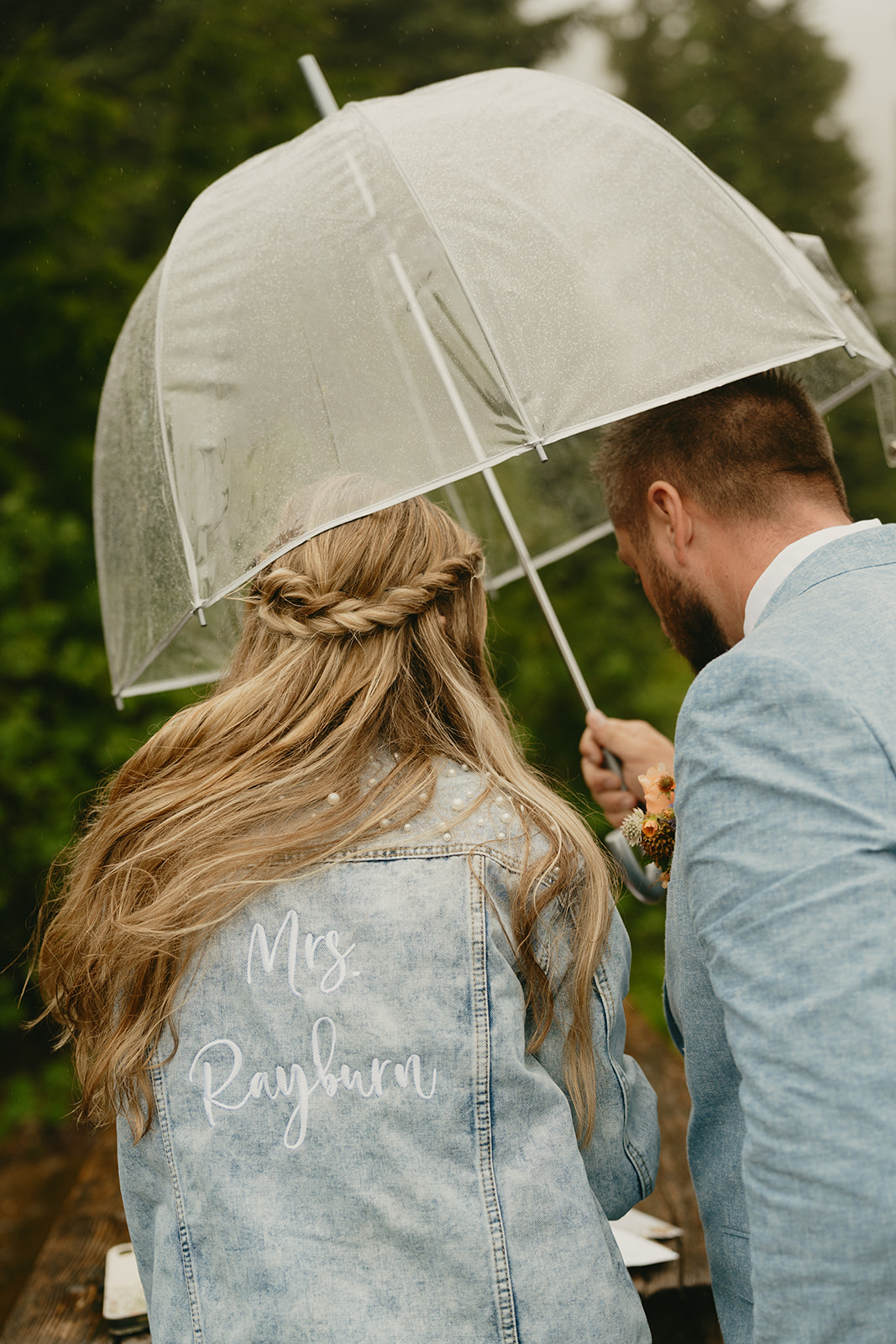 Close up of couple walking together while holding clear umbrella while bride wears personalized denim jacket