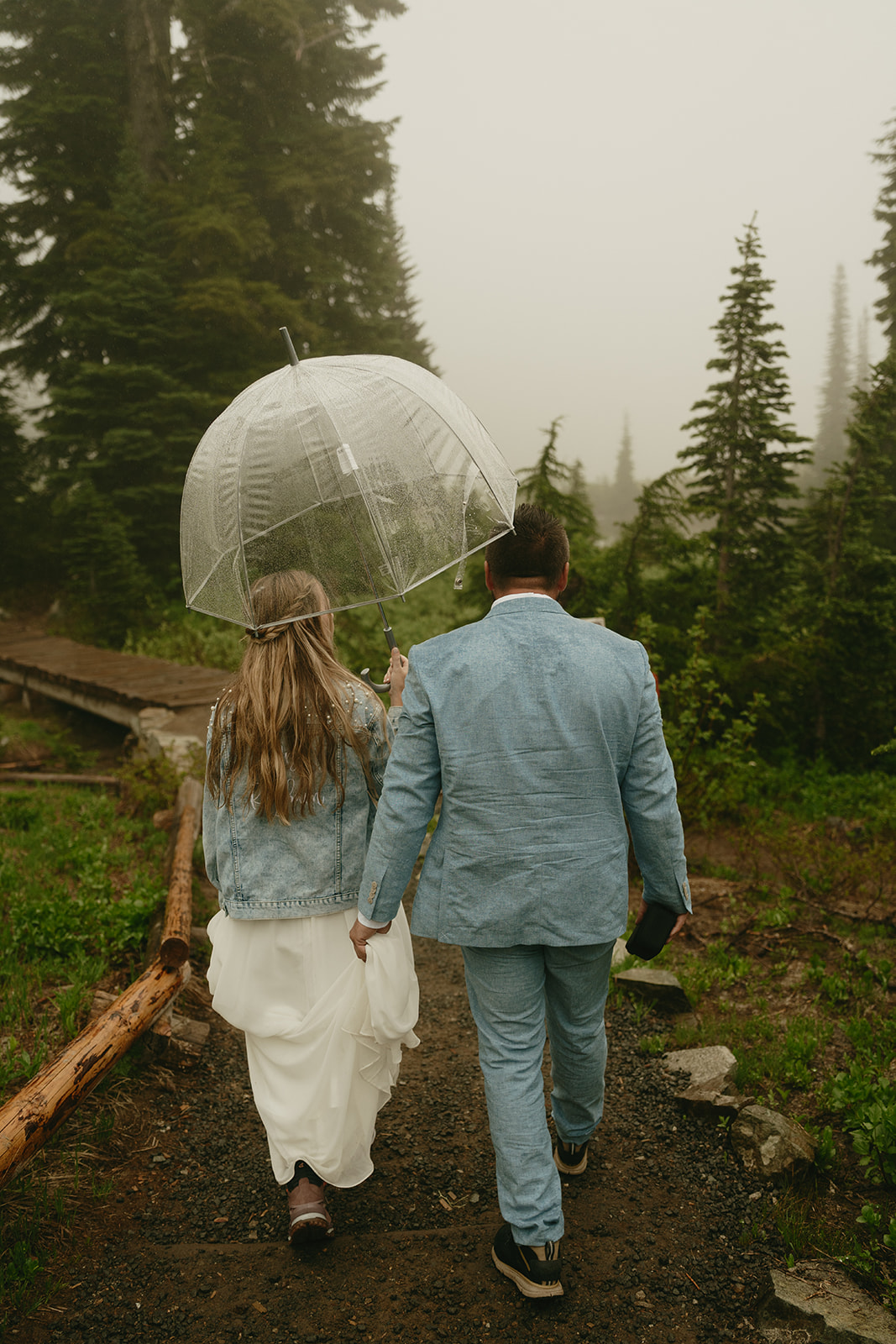Newly married couple walking together while clear umbrella as groom holds bride's wedding dress