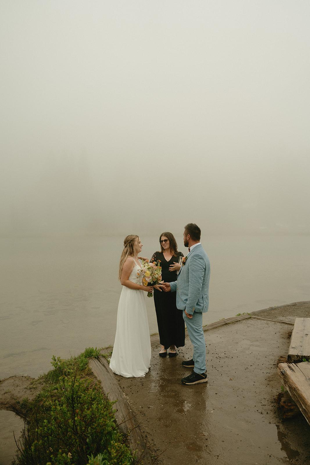 Bride and groom standing with their officiant on a foggy lakeshore, exchanging quiet vows during their elopement in Mount Rainier National Park.