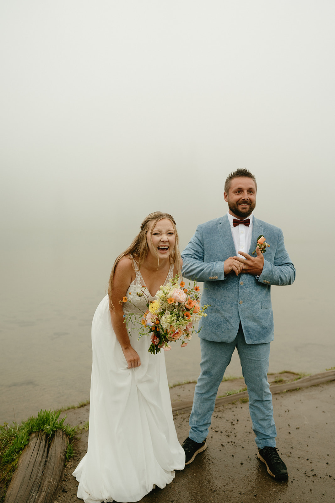 Bride and groom sharing a joyful laugh on the edge of a misty lakeshore, bouquet in hand.