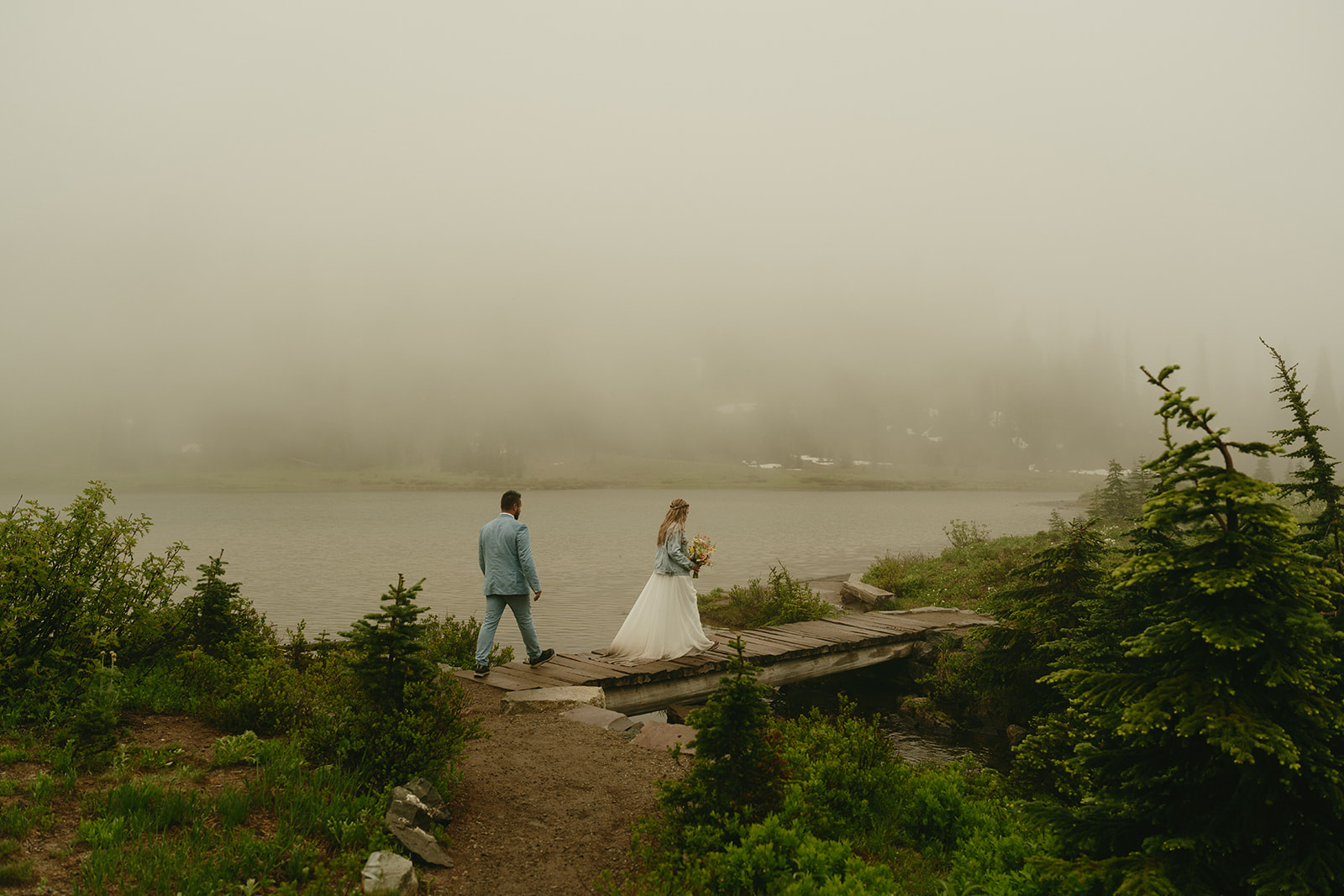 Bride and groom walking across a narrow wooden bridge near the water, surrounded by fog and alpine greenery.