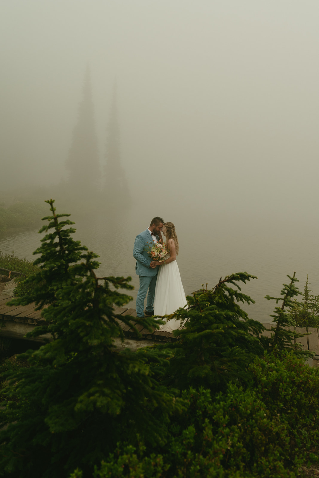 Couple standing close on a trail near a fog-covered alpine lake in Mount Rainier National Park.