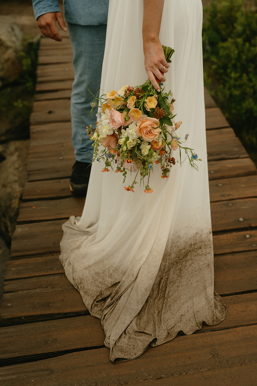 Detail shot of the bride’s mud-kissed dress hem and colorful bouquet on the wooden trail.
