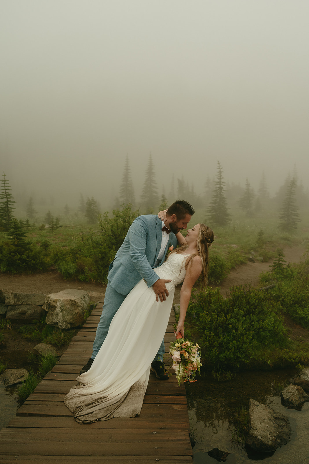 Groom gently dipping the bride for a kiss on a wooden bridge, fog and evergreens in the background.