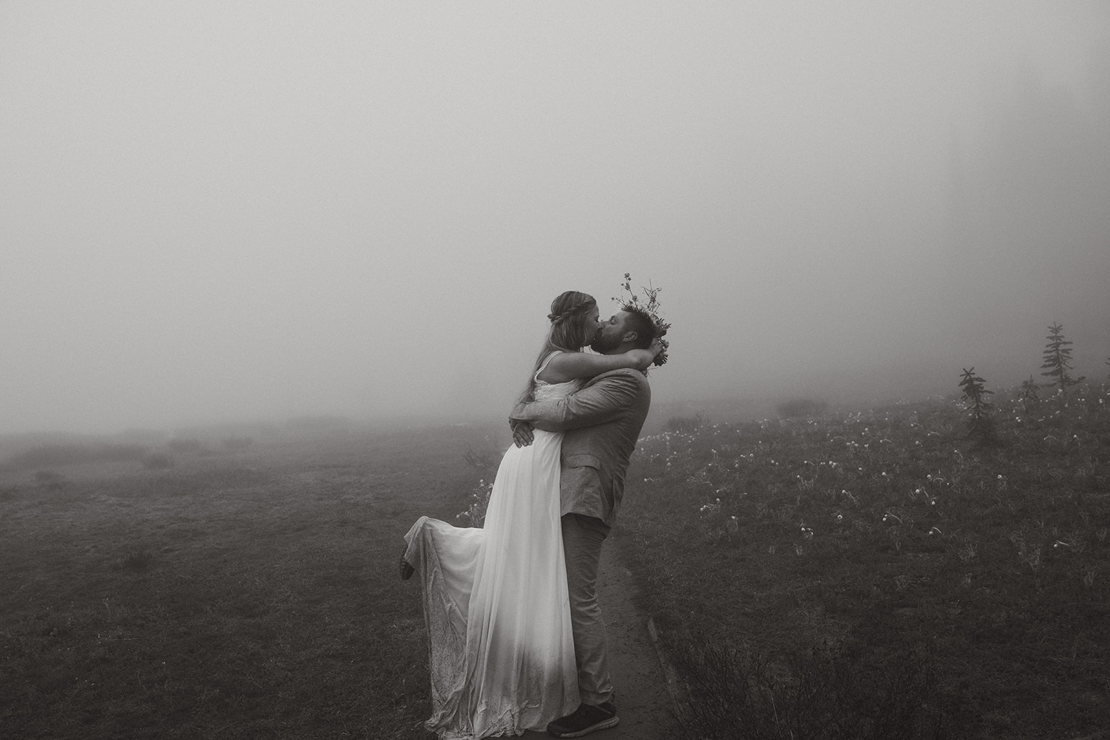 Black and white photo of a couple kissing in the mist, her dress trailing behind as he lifts her off the ground.