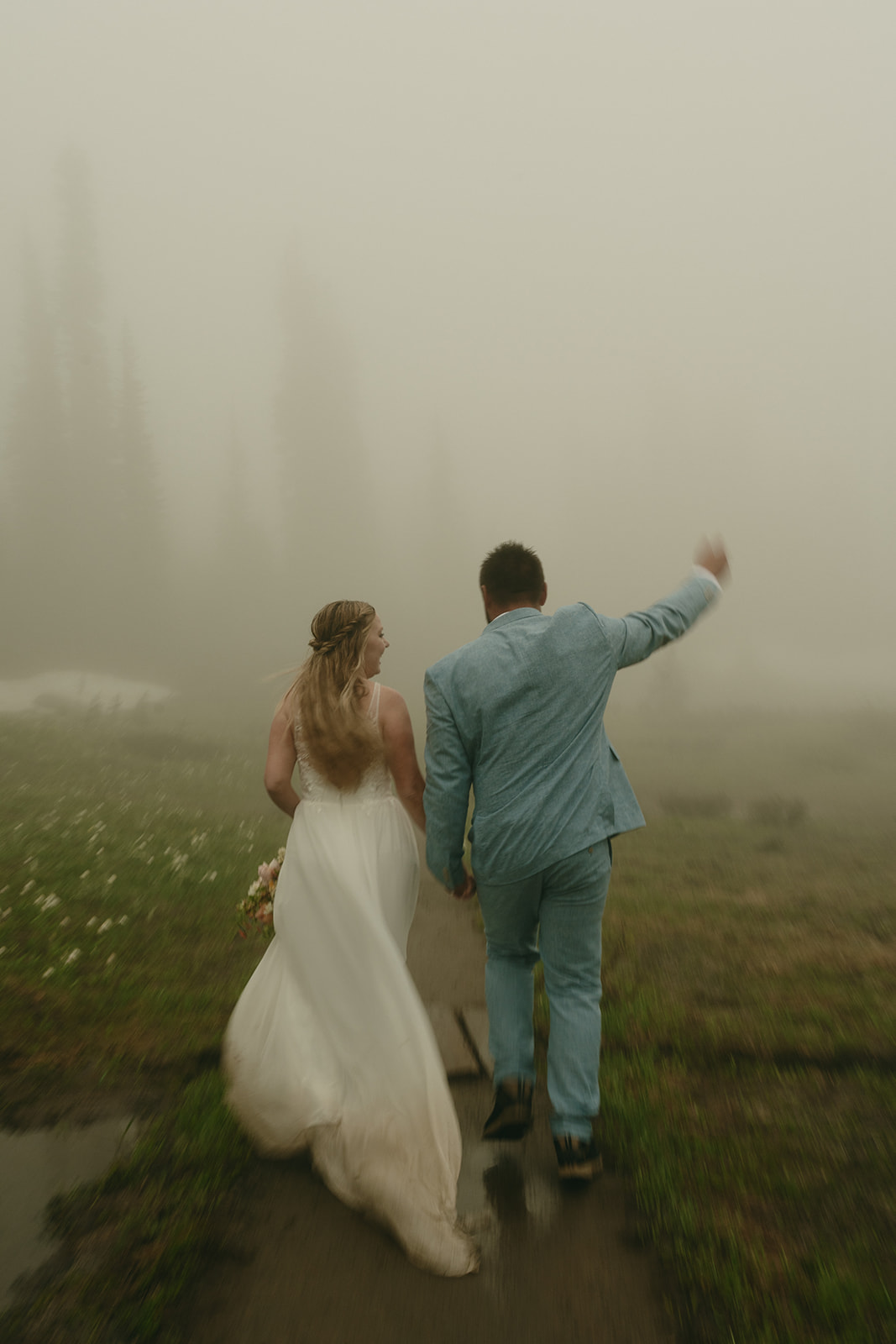 Bride and groom walking into the fog, her dress trailing behind them on a wooden path.