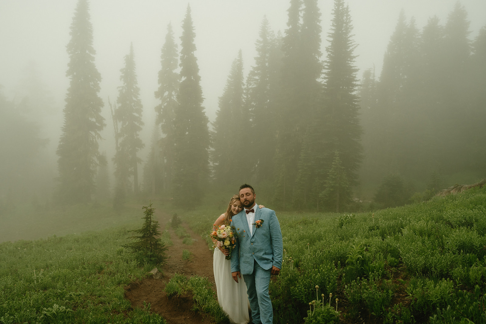Couple standing close together in a misty alpine meadow, the bride gently leaning into her partner with trees rising behind them.