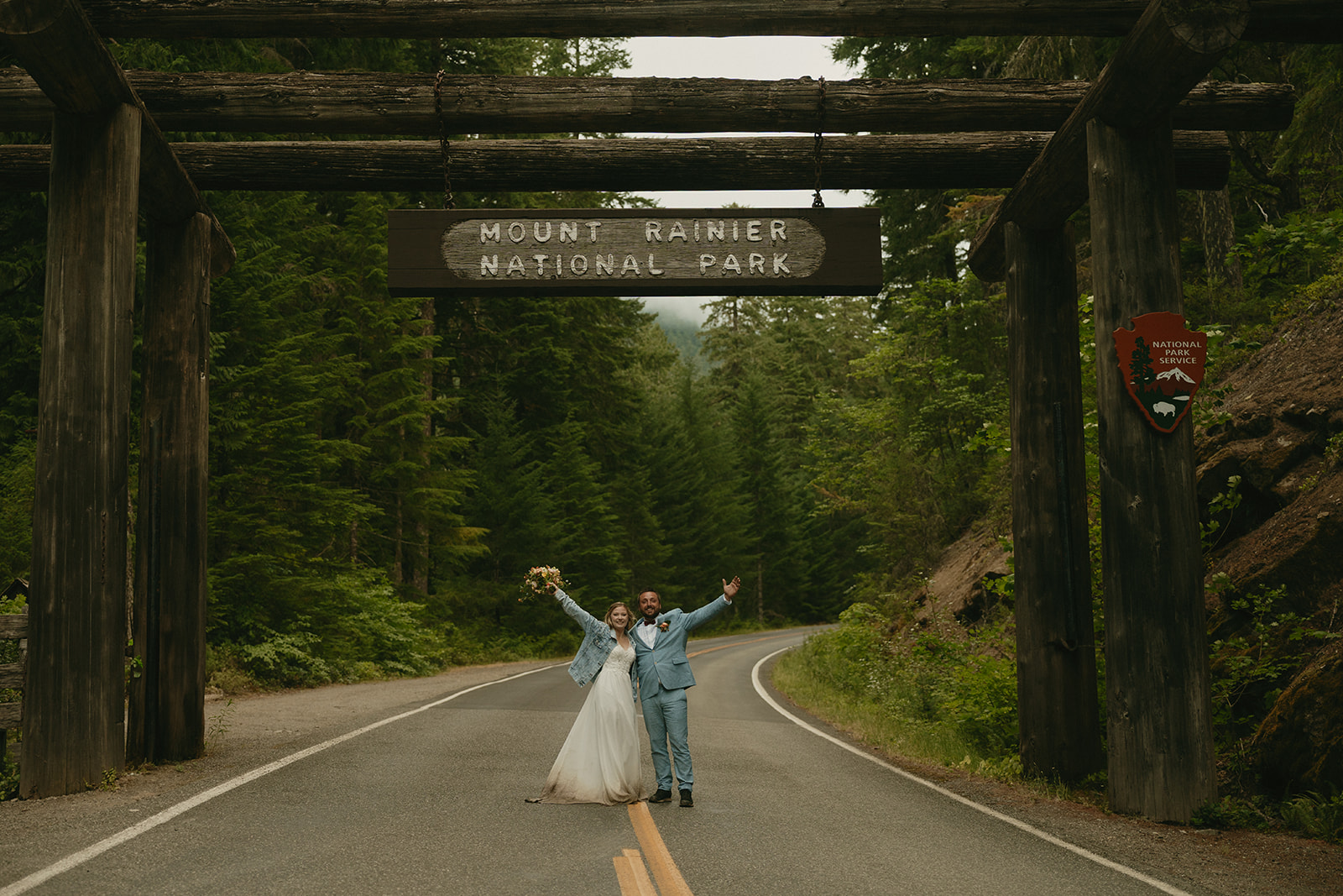 Elopement couple celebrating beneath the Mount Rainier National Park entrance sign, arms raised in joy.
