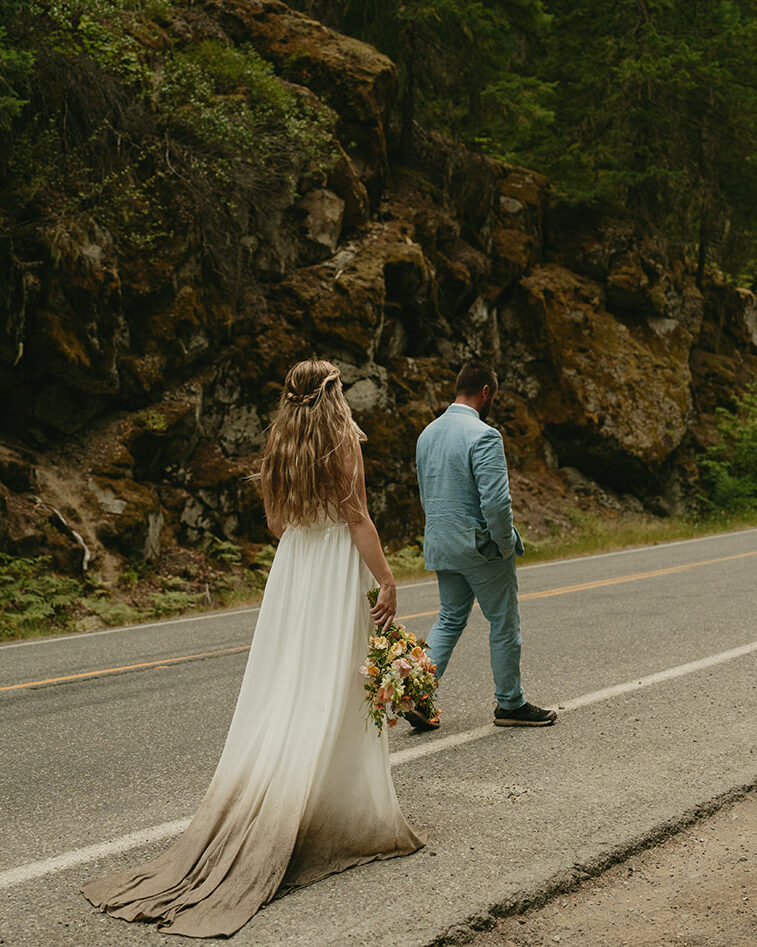 Bride and groom walking along a quiet forest road, her dress carrying traces of the earth from their elopement day.