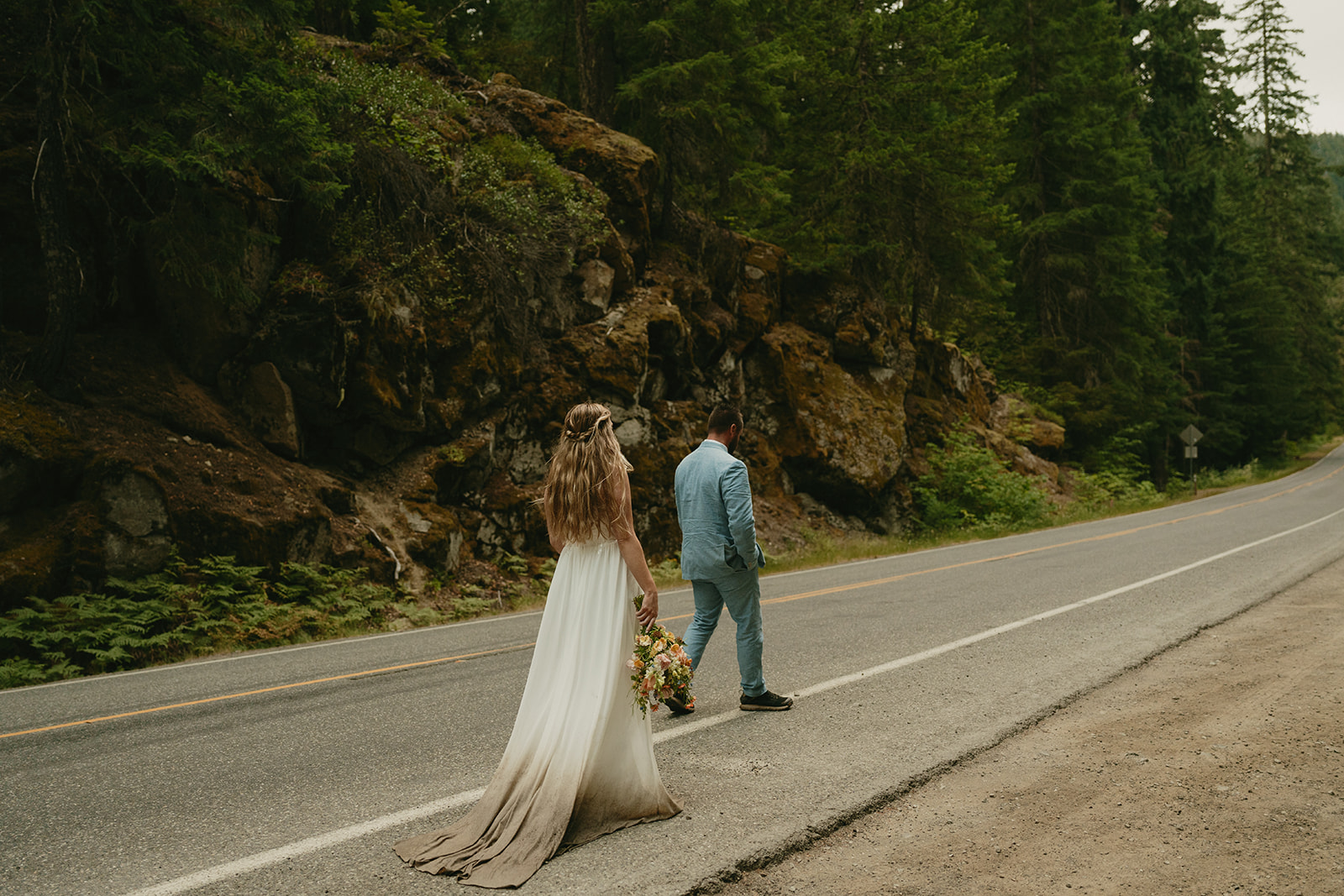Bride and groom walking along a quiet forest road, her dress carrying traces of the earth from their elopement day.