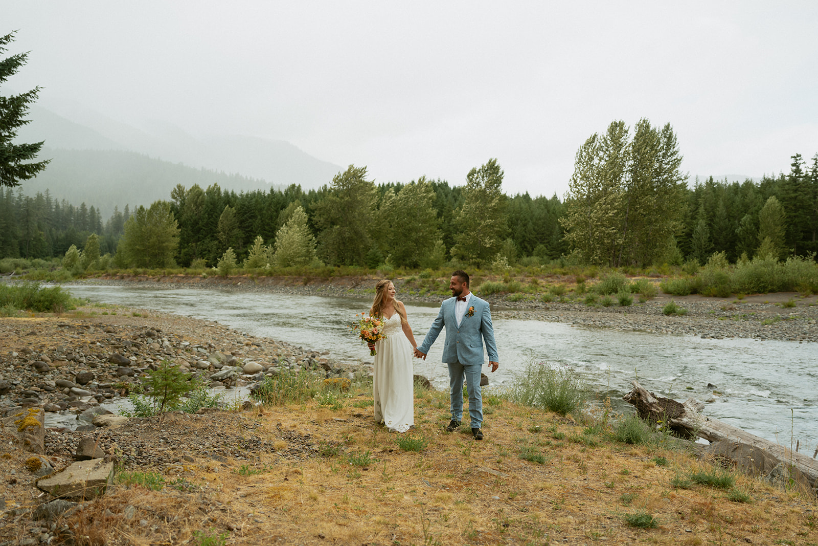 Bride and groom walking along a quiet forest road, her dress carrying traces of the earth from their elopement day.