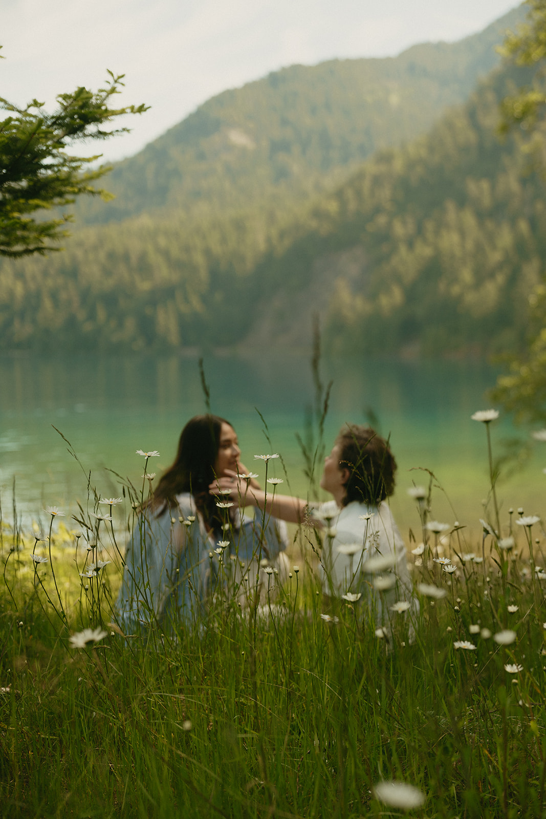 A couple sits in a wildflower meadow overlooking a quiet mountain lake, sharing a soft, playful moment surrounded by nature.