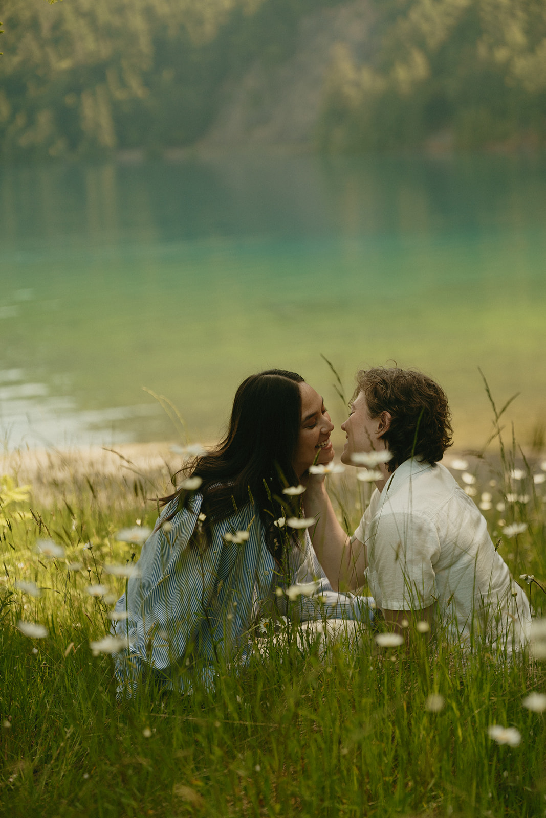 A couple sits in a meadow by a turquoise lake, leaning in for a kiss surrounded by daisies and summer light.