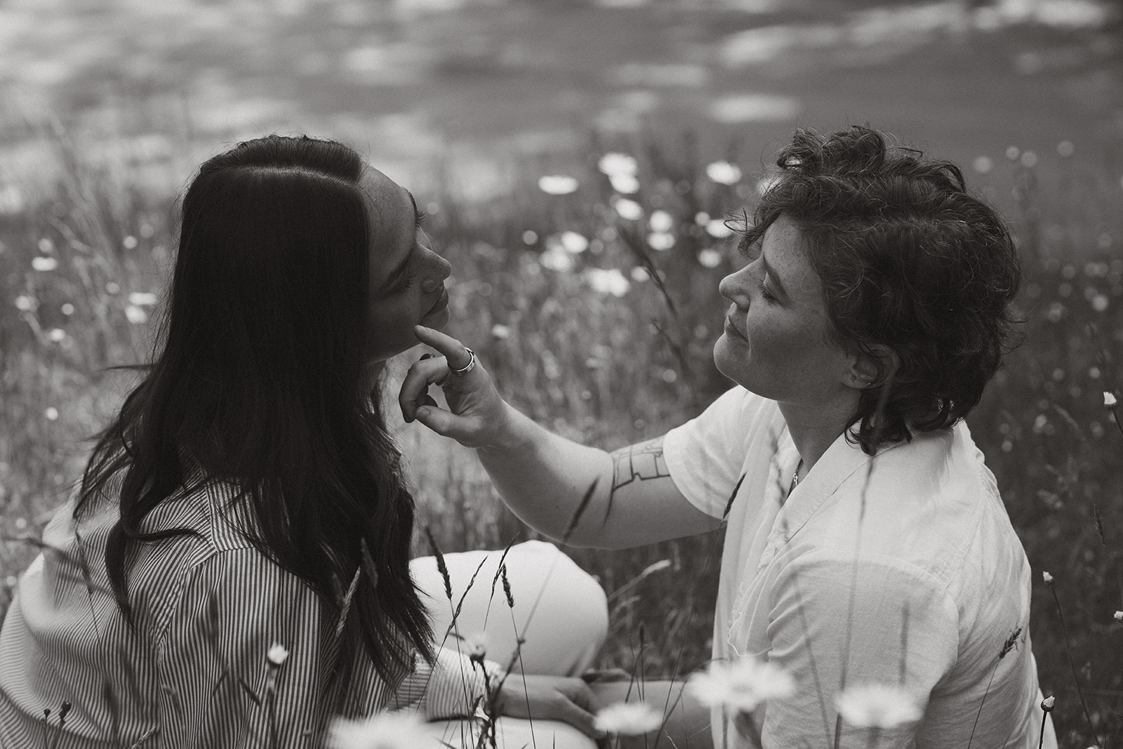 In a soft black-and-white frame, one partner gently touches the other’s chin as they sit in a wildflower-filled meadow, lost in a quiet moment together.