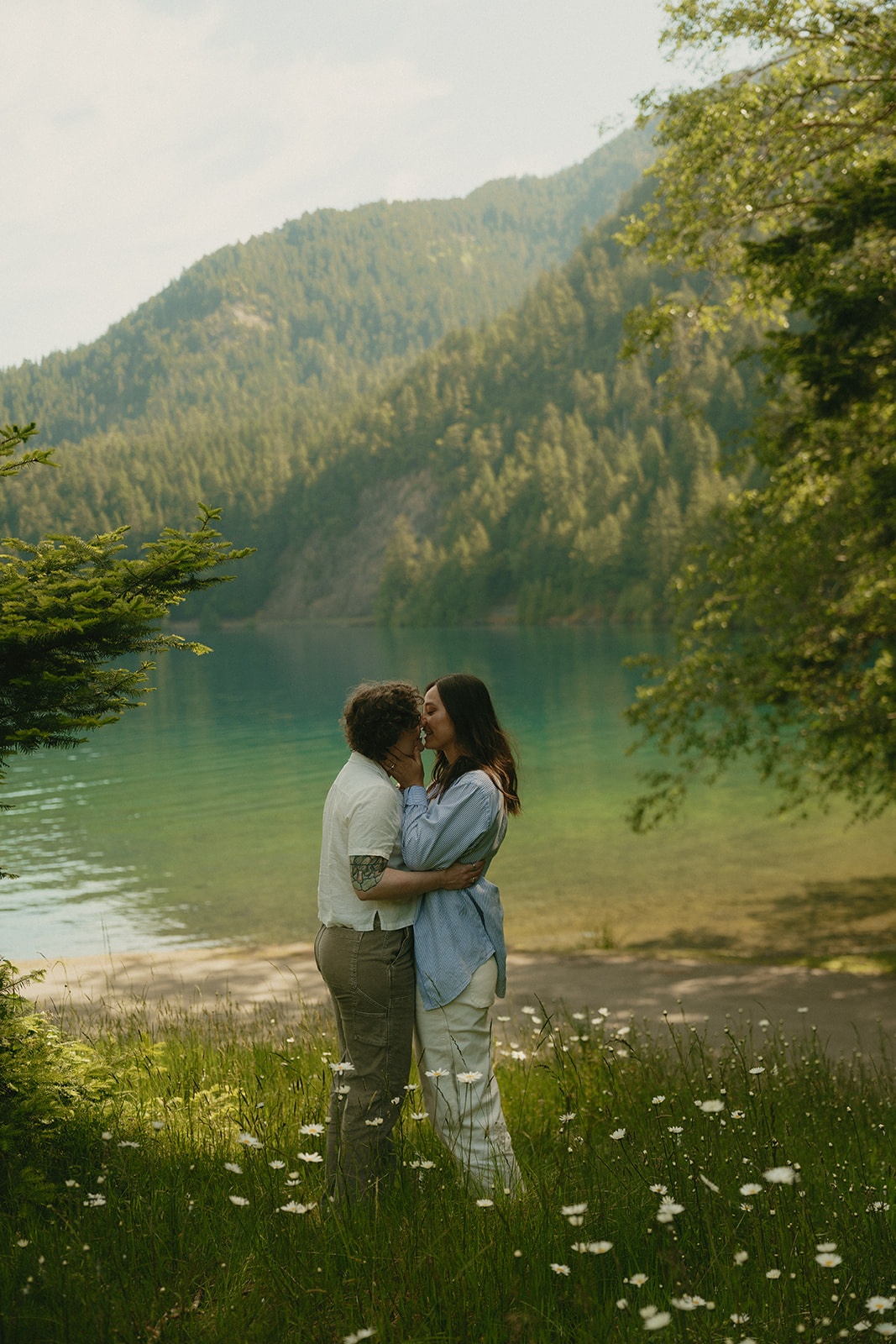 A romantic, cinematic moment as a couple embraces in a wildflower-filled meadow beside a clear alpine lake, mountain ridges rising behind them.
