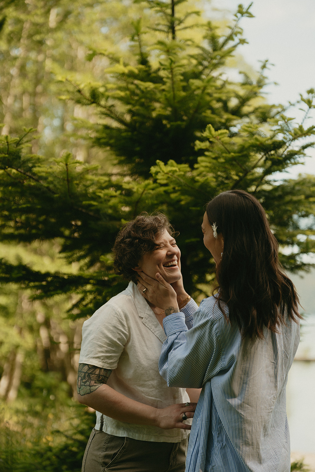 A candid moment of laughter between two partners beneath the trees, framed by evergreens and soft summer light.