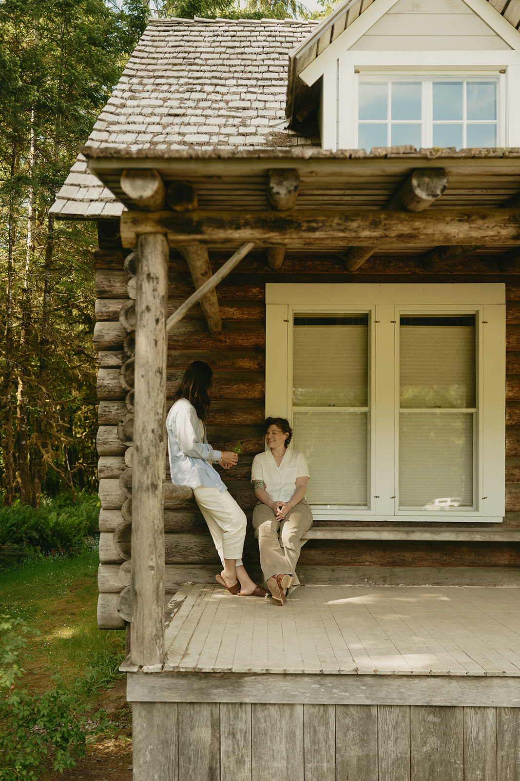 Two people sit on the porch of a cozy log cabin, sharing a quiet conversation in the stillness of the woods.