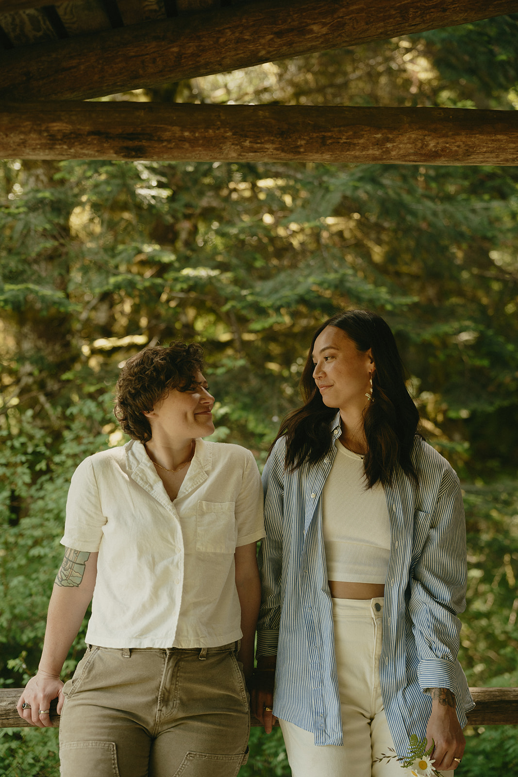 A couple holds hands beneath a wooden canopy in a quiet forest, exchanging soft smiles in dappled light.