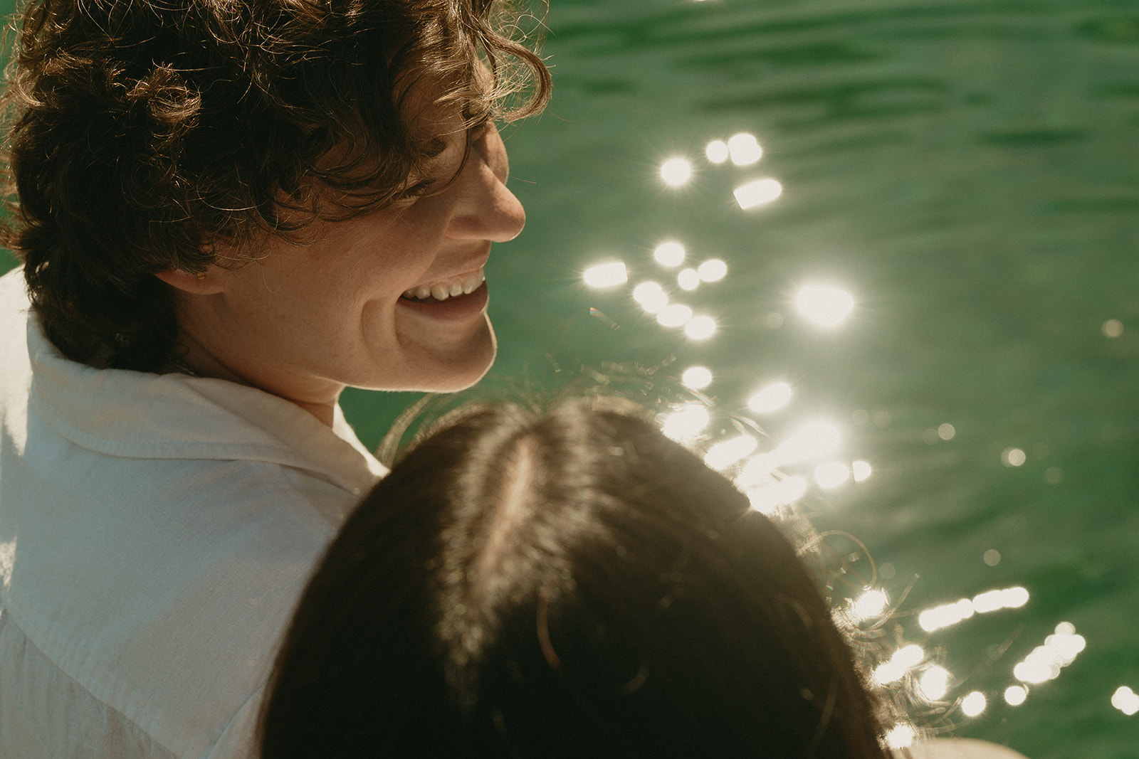 Close-up of a couple sitting at the edge of a lake, sunlight sparkling on the water as they share a quiet, joyful moment together.
