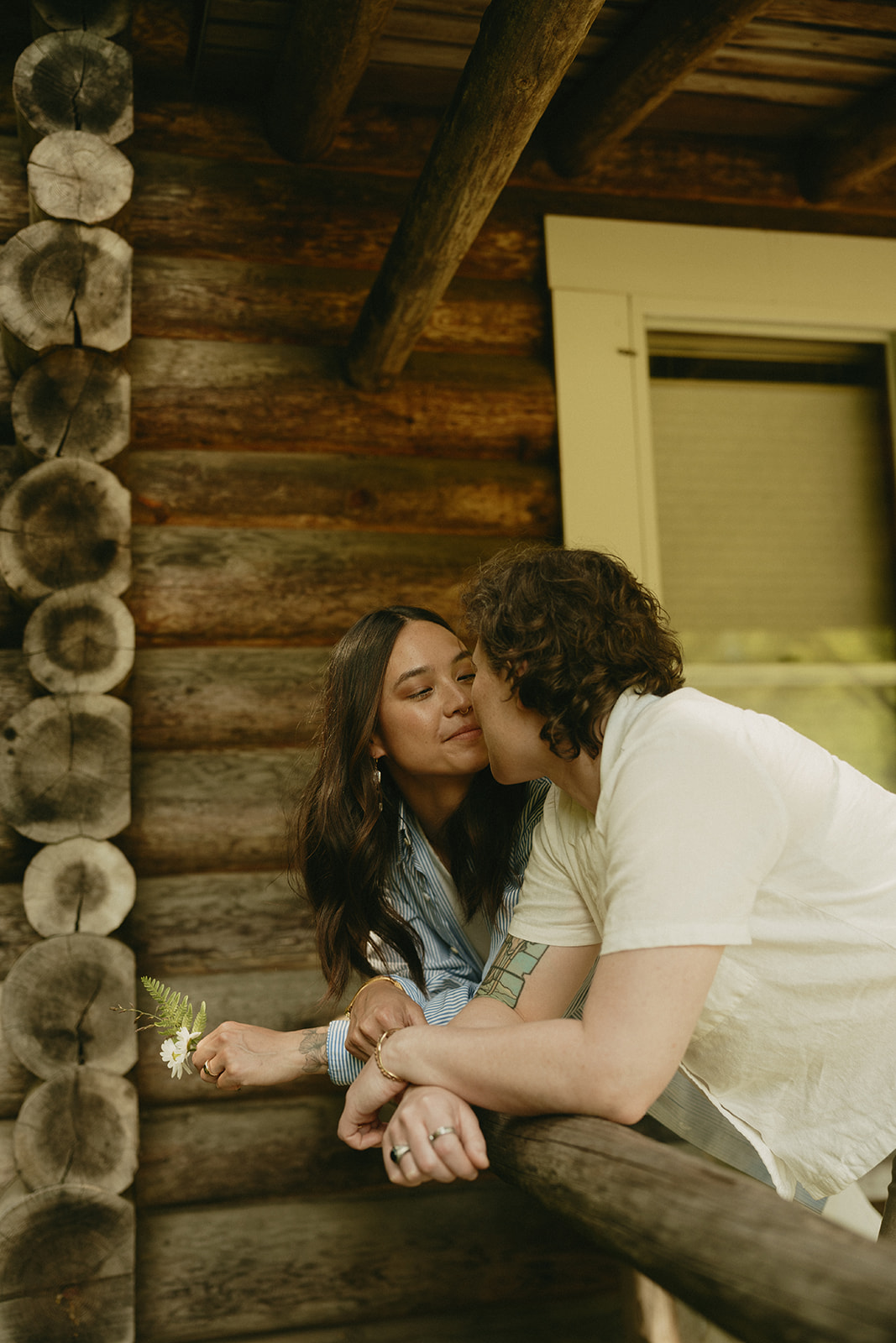 Leaning in on a rustic cabin porch, a couple shares a quiet moment, soft smiles and summer light surrounding them.