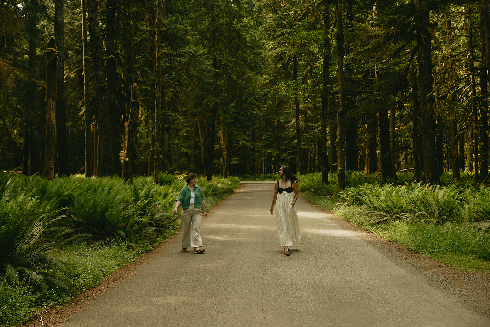 Two women walk side by side down an empty road through an old-growth forest, ferns lining the quiet path.