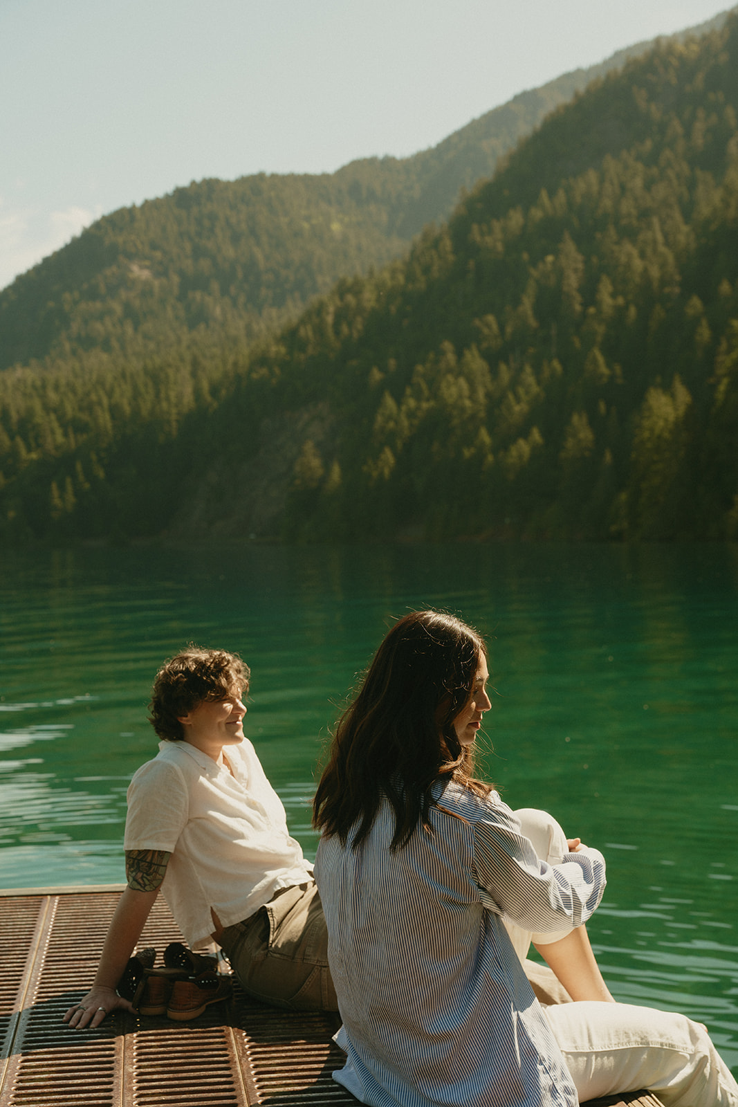 A couple sits on a quiet dock overlooking emerald-green lake water, surrounded by forested mountains and soft afternoon light.