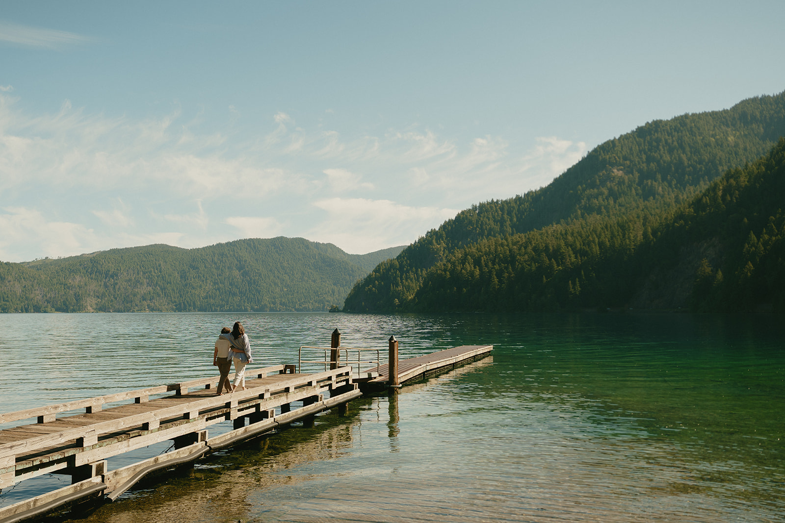 A couple walks down a weathered dock, surrounded by a vast mountain lake and still morning light.