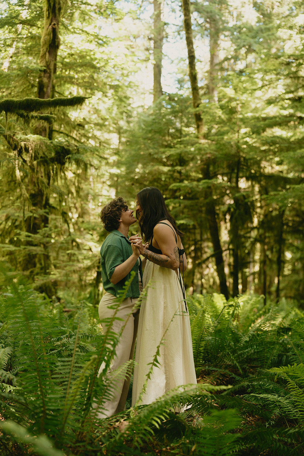 A couple stands close together in a lush fern-covered forest, sharing a quiet kiss beneath filtered sunlight.