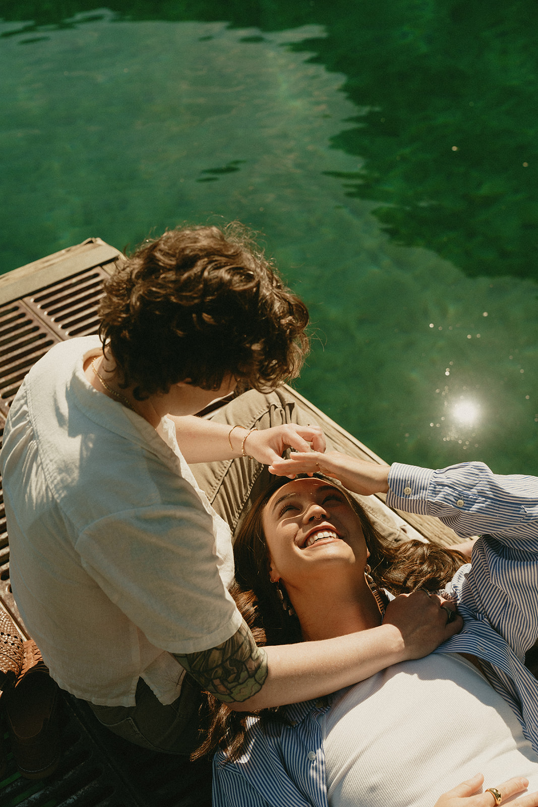 One partner lays back on the dock, smiling up at the other in a quiet lakeside moment of connection.