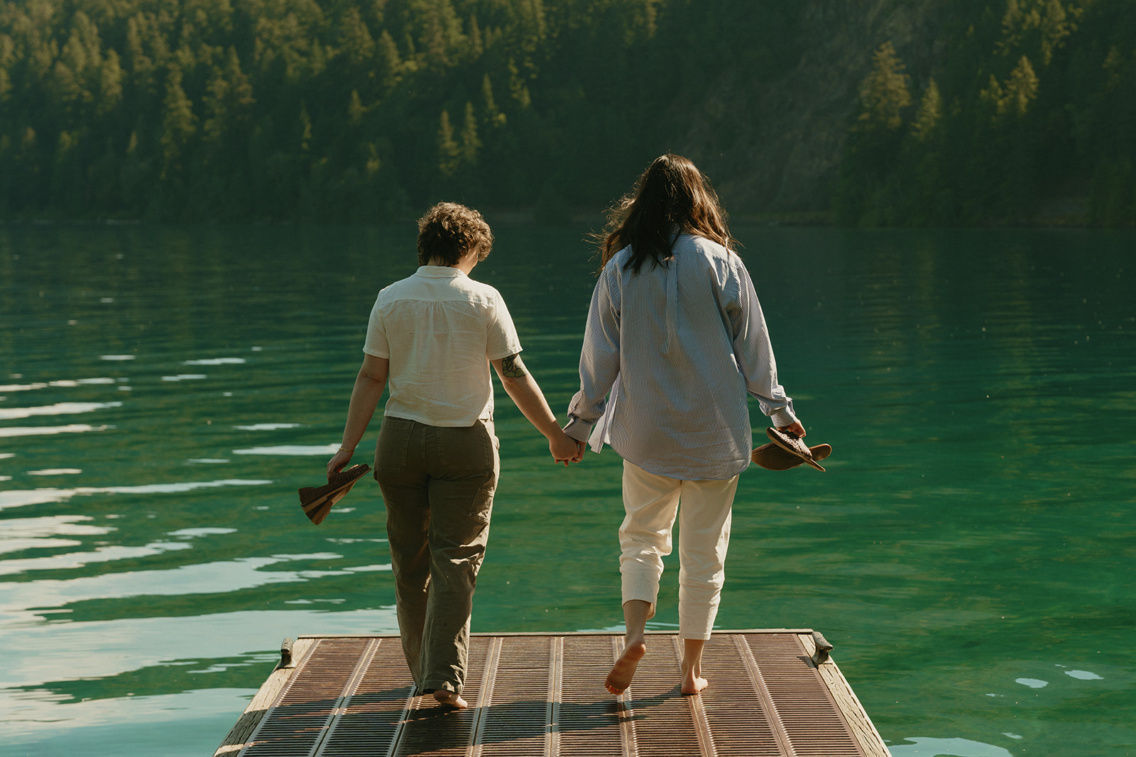 Walking barefoot on a lakeside dock, a couple holds hands and carries their shoes, surrounded by still green water.