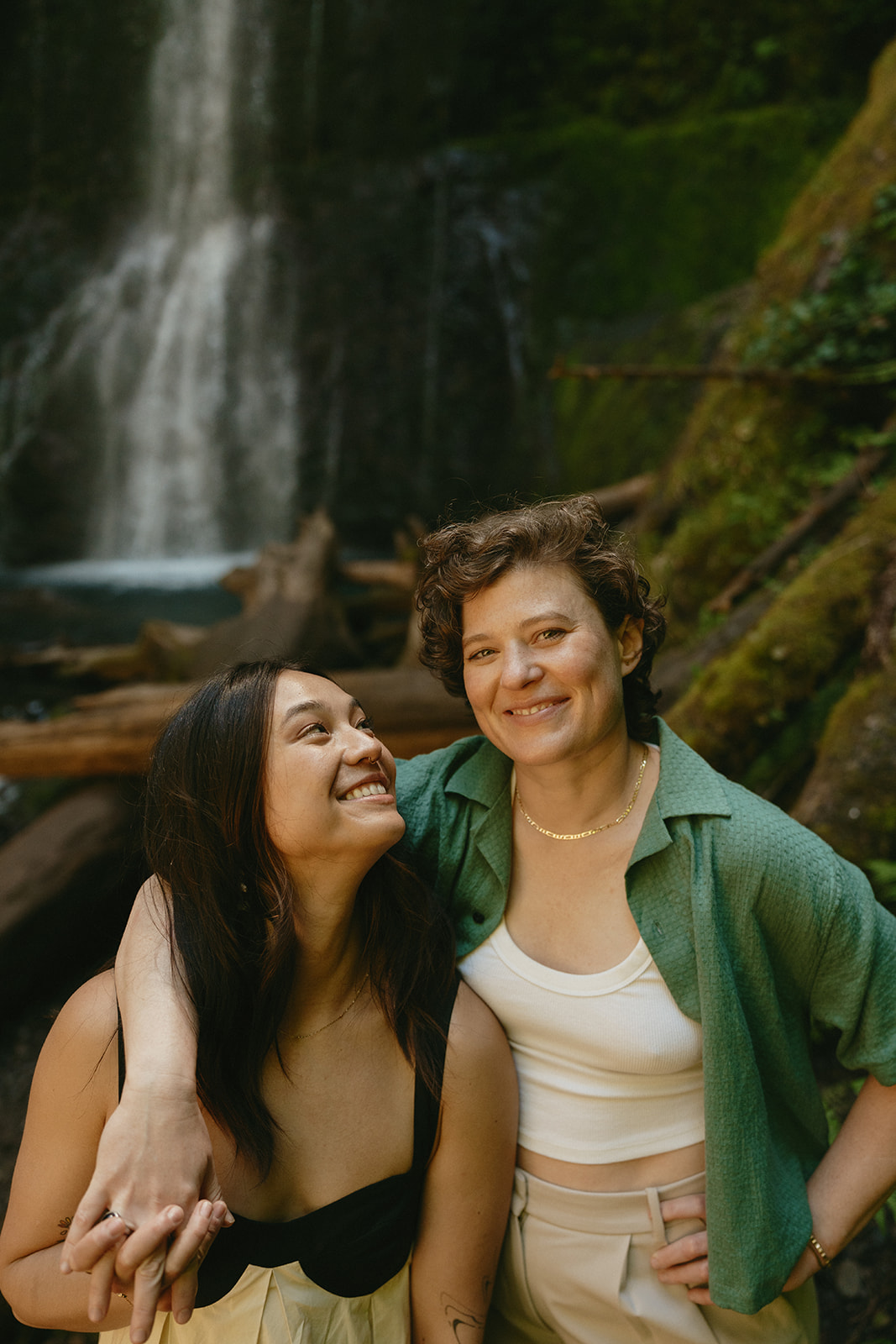 A joyful portrait of two women in front of a cascading waterfall, one smiling at the camera while the other looks up with love.