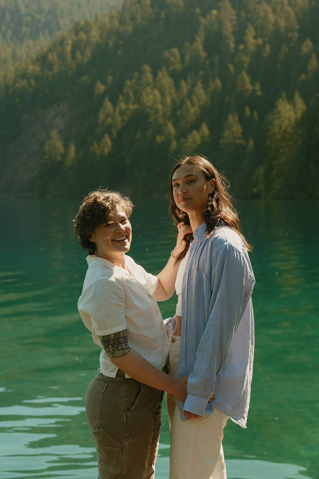 A candid portrait of a couple standing close by the water, one smiling toward the camera as golden light reflects off the lake.