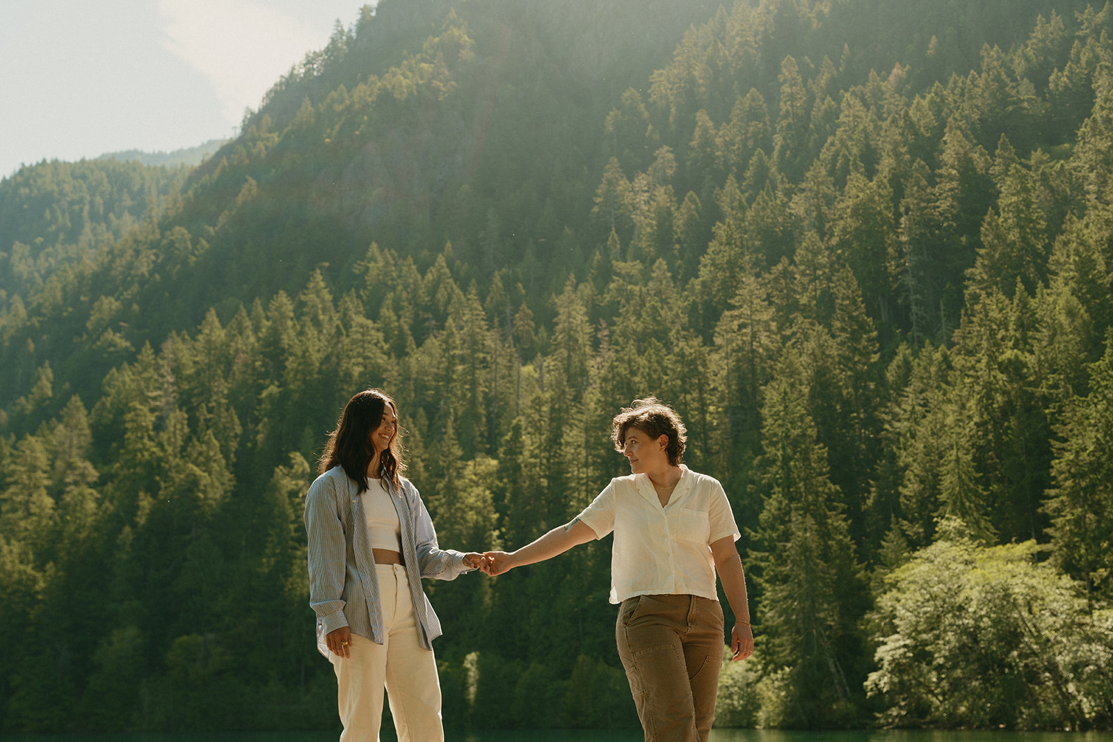 Holding hands against a backdrop of towering evergreens and late afternoon sun, a couple walks along the lake’s edge together.