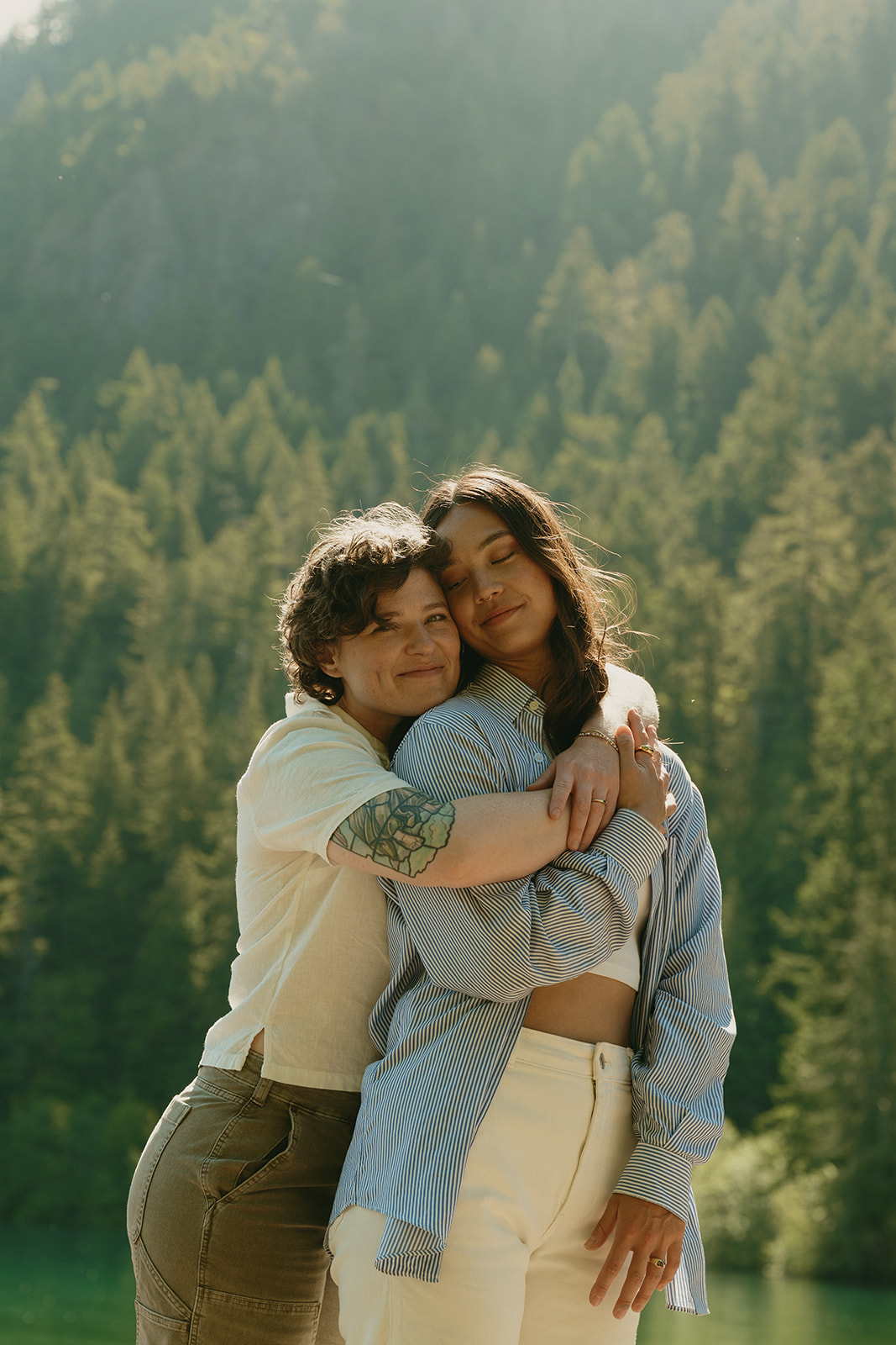 One partner wraps the other in a soft embrace, their eyes closed in the golden light by the lake.