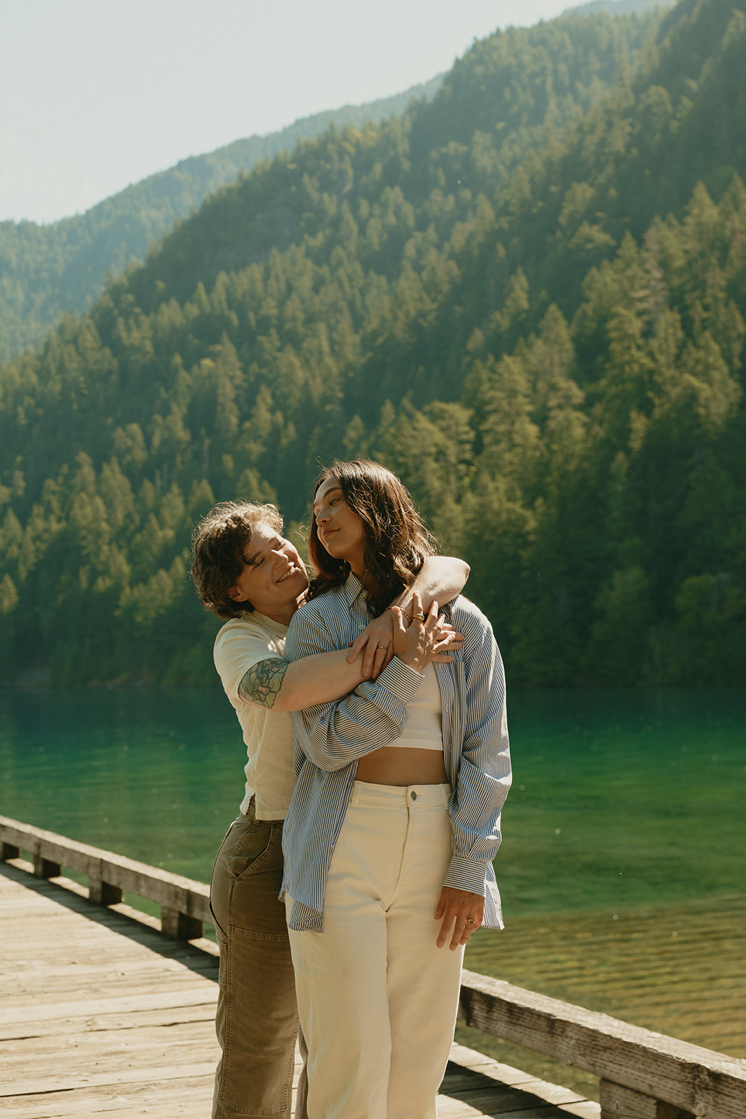 Captured in a lakeside hug, one partner rests their arms around the other’s shoulders as they stand side by side on a wooden dock.