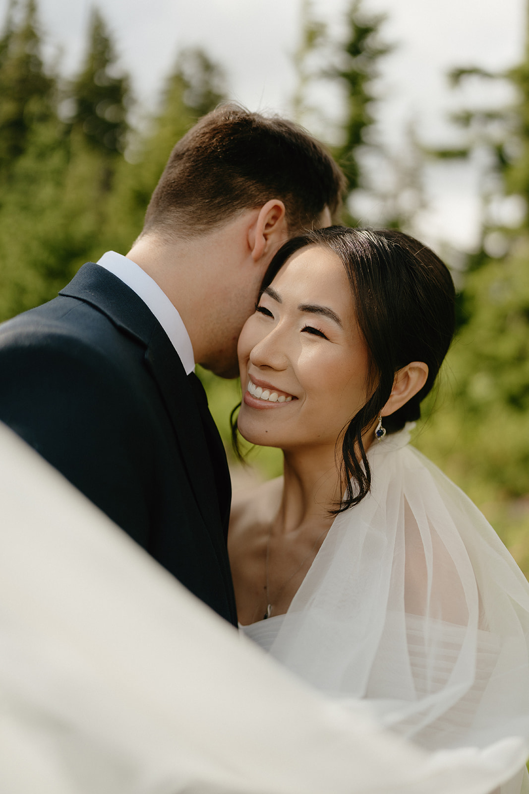 Bride smiles softly as the groom holds her close, veil flowing in a quiet forest setting.